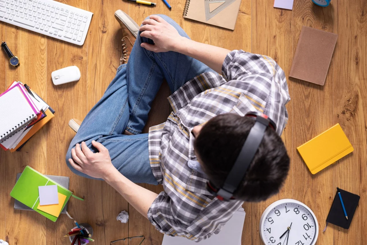 اهمال کاری در درس خواندن؛ عوامل ایجادکننده و مقابله با آن_اسپایدر 3 پسربچه نشسته روی زمین چوبی با هدفون روی سر و دفترچه ها و ساعت روی زمین